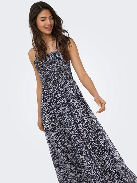 Vrouw met lange, golvende bruine haren, draagt een marineblauwe maxi jurk met spaghettibandjes en een witte geometrische print.