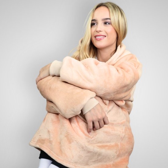 Een vrouw met blond haar draagt een oversized beige hoodie deken. Ze zit, omhelst zichzelf en kijkt naar rechts.