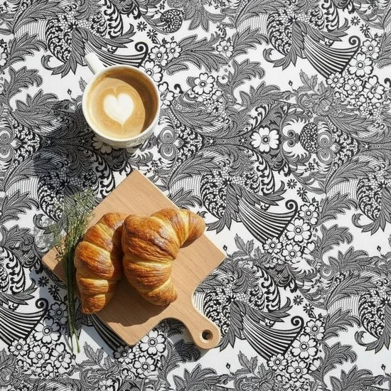 Zwart-wit bloemenpatroon tafelkleed met koffiekopje en croissants op een houten plank.