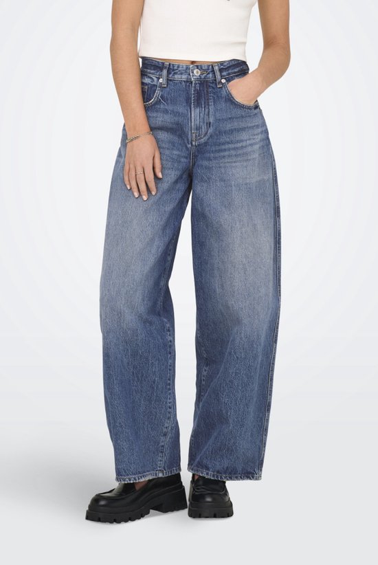 Model in wijde, middendonkere spijkerbroek met hoge taille en vier zakken. Zwarte schoenen.