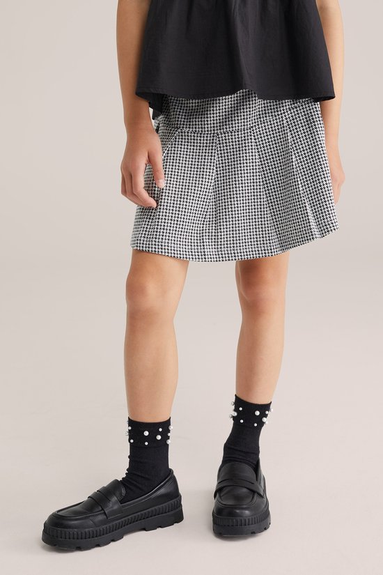 Meisje draagt een geruite rok, zwarte sokken met witte stippen en zwarte loafers.