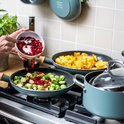 Een hand voegt granaatappelpitjes toe aan spruitjes in een koekenpan, naast aardappelen in een andere pan op een gasfornuis.