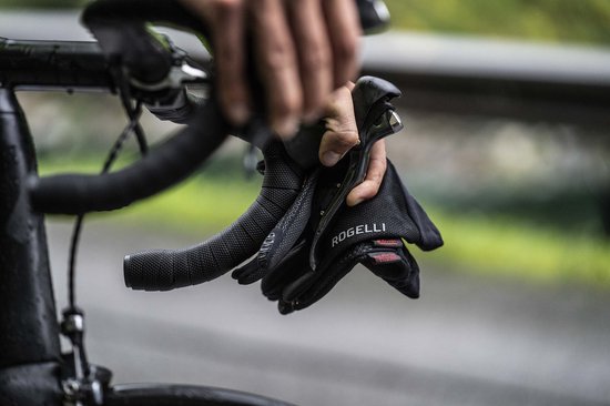 Een persoon houdt een paar zwarte Rogelli fietshandschoenen vast aan het stuur van een racefiets.