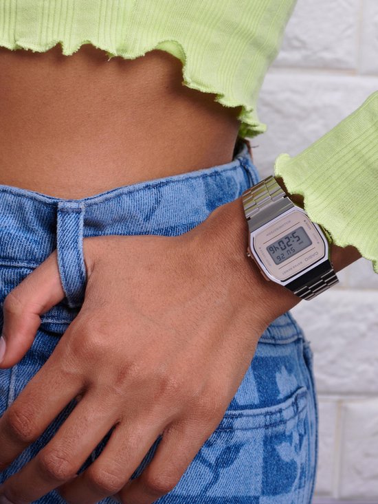 Zilverkleurig Casio horloge op de pols van een vrouw, gedragen met een felgroene top en gebloemde jeans.