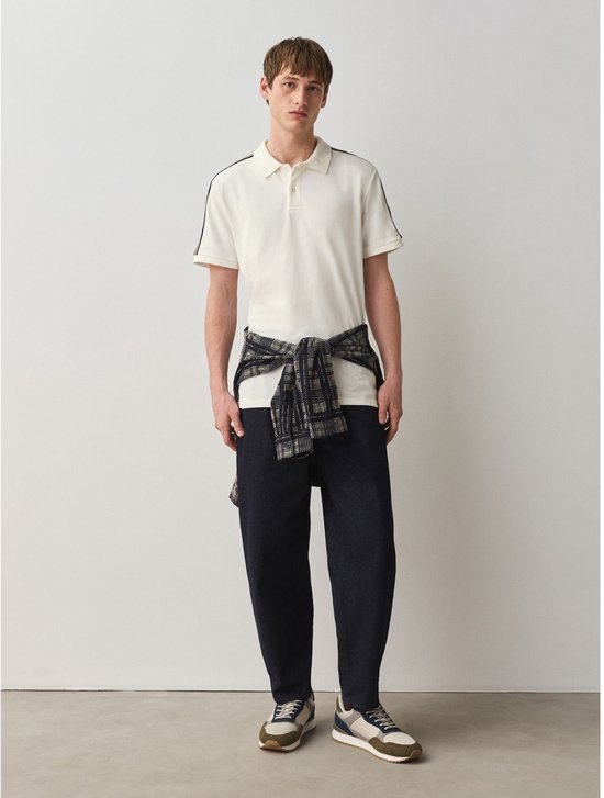 Man draagt een crème poloshirt met strepen op de mouwen, met een geruit vest om zijn middel gebonden.