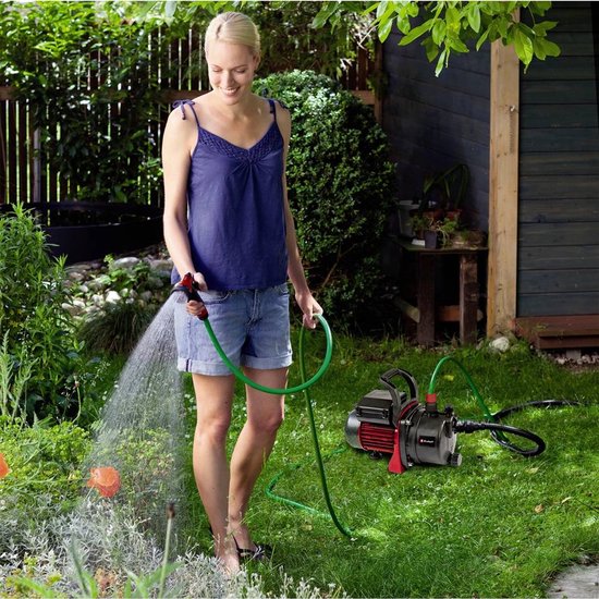 Vrouw met blauw topje en spijkerbroek sproeit tuin met tuinslang, naast een zwarte pomp met rode details.