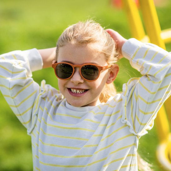 Jeune fille souriante aux cheveux blonds, portant des lunettes de soleil orange pour enfants et un pull rayé, mains dans les cheveux.