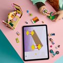Kind bouwt Lego Friends speelgoedhuisje, met tablet en accessoires op een roze tafel.