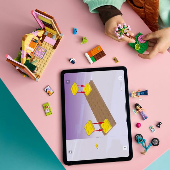 Kind bouwt Lego Friends speelgoedhuisje, met tablet en accessoires op een roze tafel.