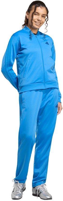 Model in blauw adidas trainingspak, bestaande uit een jack met logo en gestreepte mouwen, en een trainingsbroek. Staat met handen achter de rug.