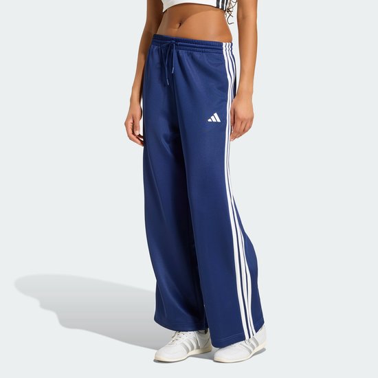 Model in een donkerblauwe wijde trainingsbroek met witte strepen op de zijkanten en witte sneakers.