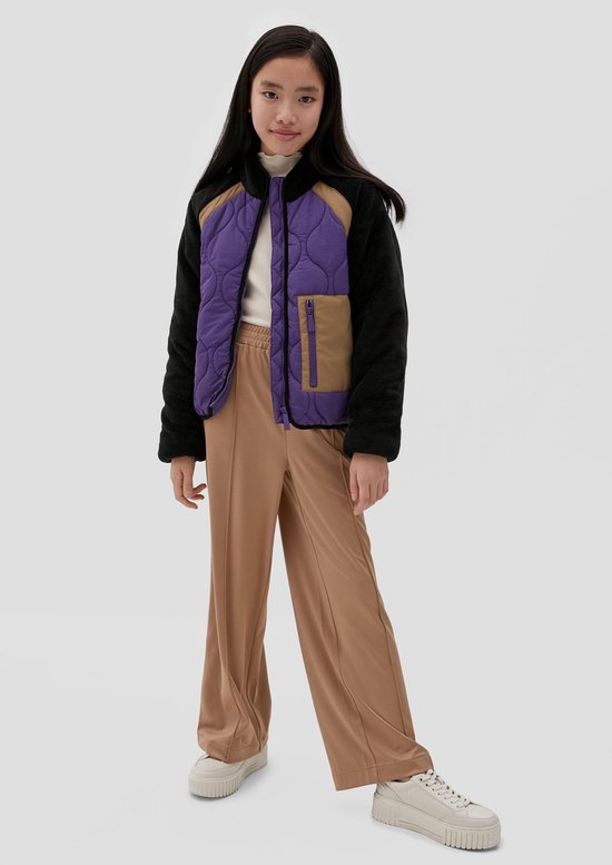 Model draagt een paars en bruin gewatteerd jack met zwarte fleece mouwen en een beige broek.