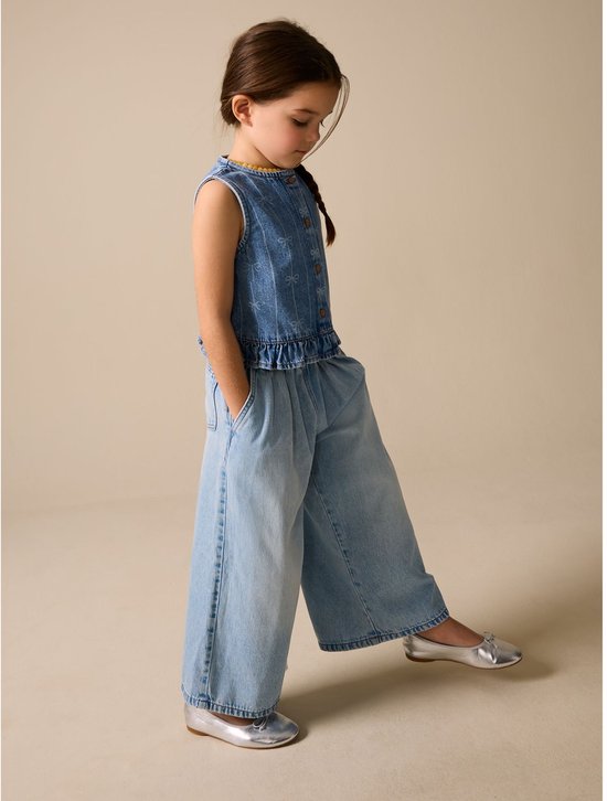 Meisje met gebloemde vlecht, draagt een mouwloos denim gilet met strikprint en wijde spijkerbroek.