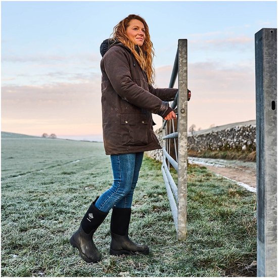 Vrouw met krullend haar, staand in een vochtig grasveld bij zonsopgang, draagt zwarte Muck Boot laarzen.