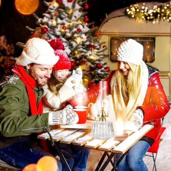 Familie geniet van winterse buitenlucht met olielamp op tafel, kerstboom en caravan op achtergrond.