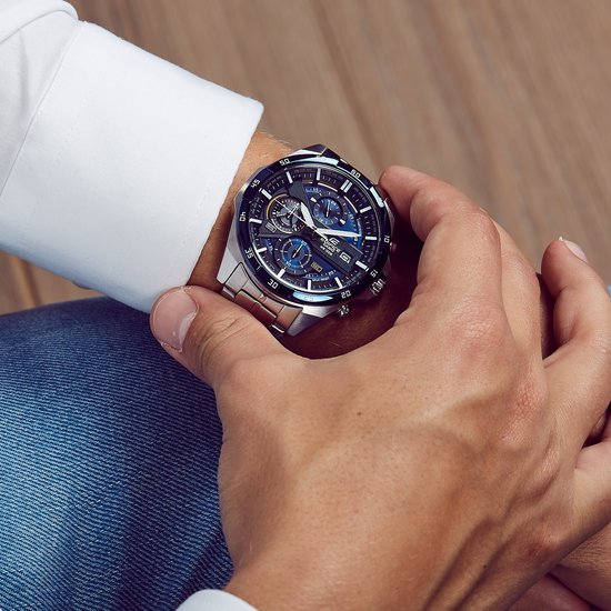 Man in wit overhemd en spijkerbroek bekijkt zilverkleurig Edifice horloge met blauwe wijzerplaat.