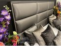 Grijze, gestoffeerde boxspring met een hoog hoofdbord en diverse sierkussens.
