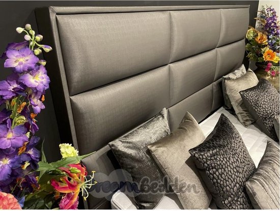 Grijze, gestoffeerde boxspring met een hoog hoofdbord en diverse sierkussens.