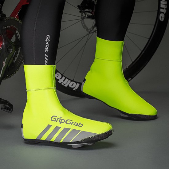 Voeten van een fietser met neongele fietsoverschoenen met reflecterende strepen, met fiets op de achtergrond.