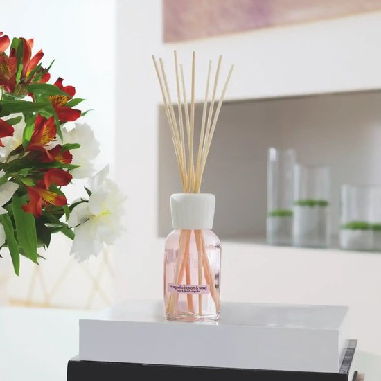 Glazen flesje met roze geurvloeistof en houten stokjes, met label 'magnolia blossom & wood'.