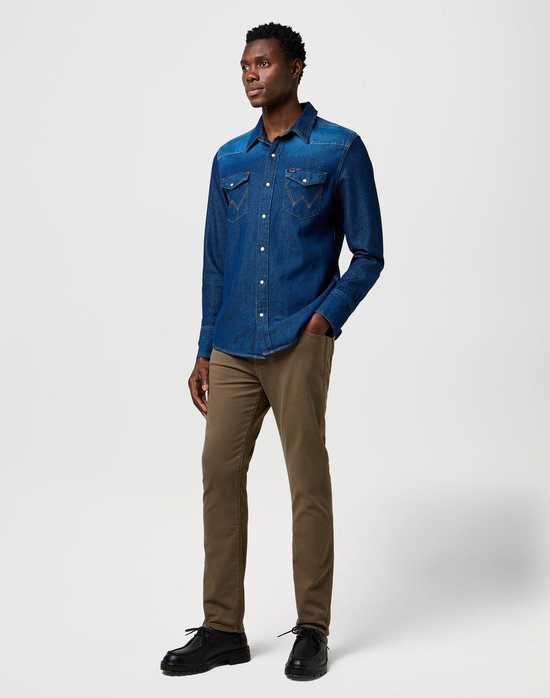 Man in olijfgroene Wrangler broek en denim blouse, staande met de hand in de zak.