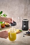 Persoon voegt rietje toe aan een glas met groene smoothie, met blender en fruit op de achtergrond.