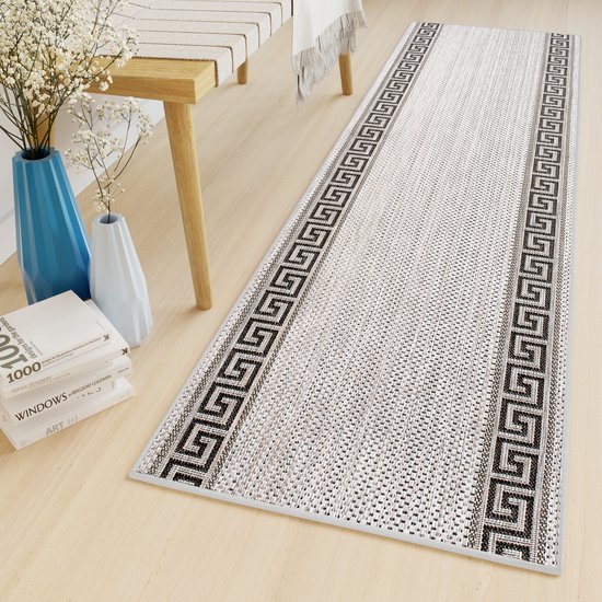Un tapis gris clair avec des bordures grecques noires dans un cadre domestique, avec des meubles et des vases.
