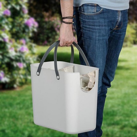 Man draagt een witte afvalbak met grijze handvatten, gevuld met papier, in een tuin.