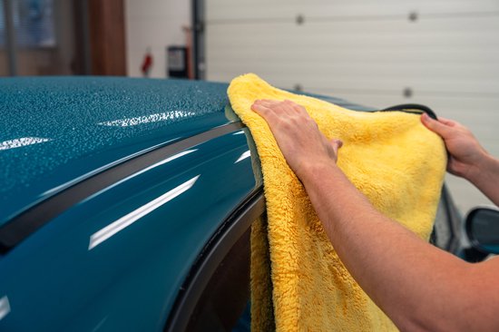 Een persoon die een felgeleende microvezel droogdoek gebruikt om de teal blauwe auto, met waterdruppels, af te drogen.