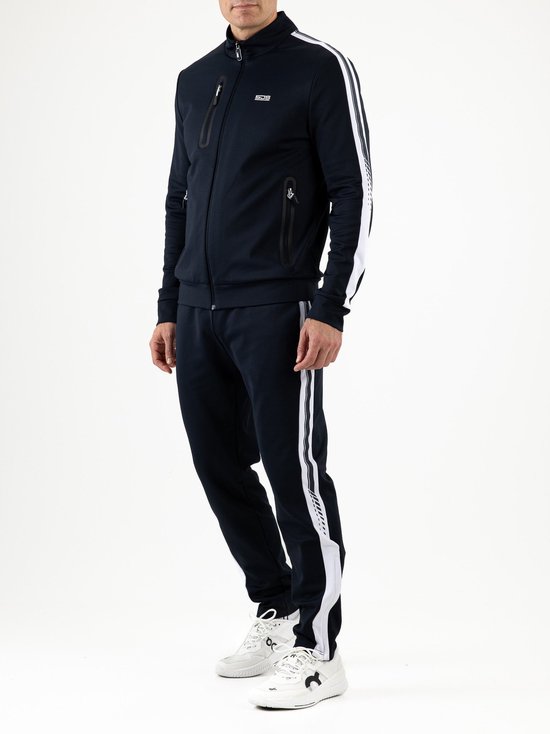 Model draagt marineblauw Sjeng Sports trainingspak met witte zijstrepen en ritszakken.