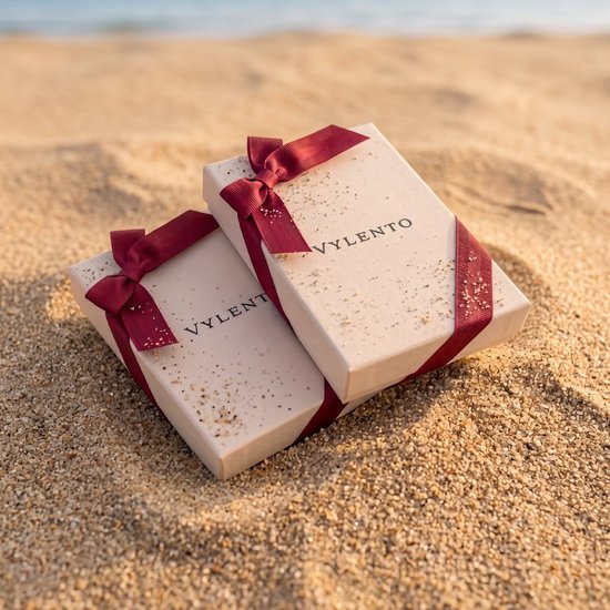 Twee beige Vylento cadeauverpakkingen met rode linten, geplaatst op zand.