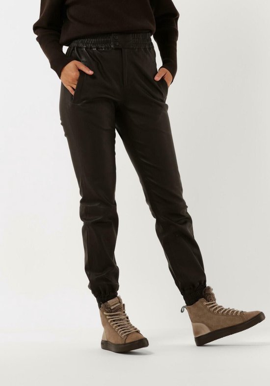 Een model draagt een bruine broek met hoge taille en elastische band. De broek heeft een leren look en is afgewerkt met elastische boorden aan de onderkant.