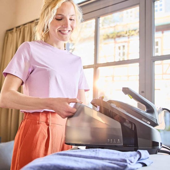 Vrouw met blonde krullen in een lichtroze shirt en oranje broek, vult het waterreservoir van een zwart stoomstrijkijzer.