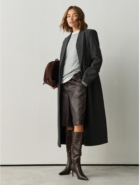 Model in een grijze jas, bruine lederen short en bruine knielaarzen met krokodillenmotief.