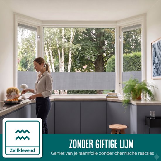 Vrouw kookt in een keuken met grijze kastjes en een erkerraam met horizontale grijze vouwgordijnen. "Zelfklevend".