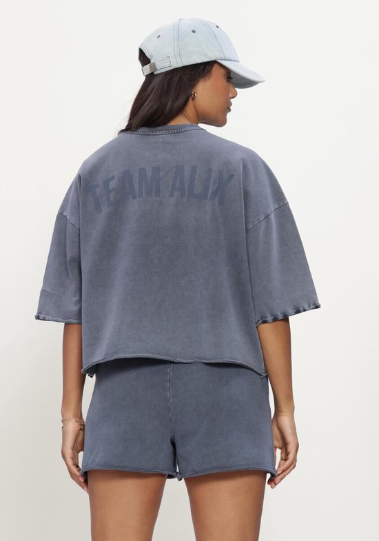 Model draagt een blauwe geknipte top met korte mouwen, gezien van de achterkant met een pet.