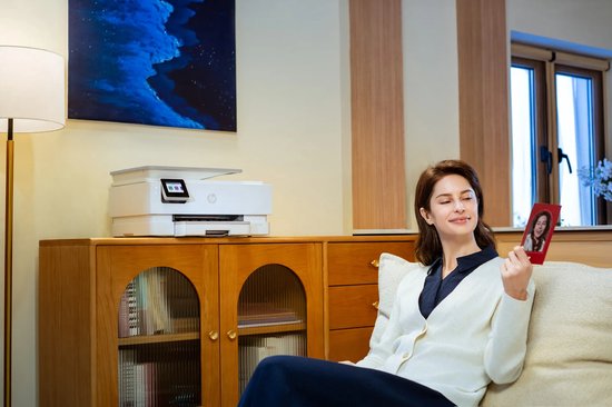 Vrouw in beige vest bekijkt foto afgedrukt door een witte HP Envy Photo printer op een houten kast.