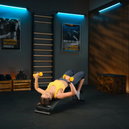 Vrouw in geel sport-top op trainingsbank met benen omhoog, handen achter hoofd.