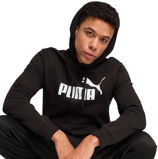 Close-up van een man zittend, die een zwarte PUMA hoodie draagt met een wit PUMA logo.