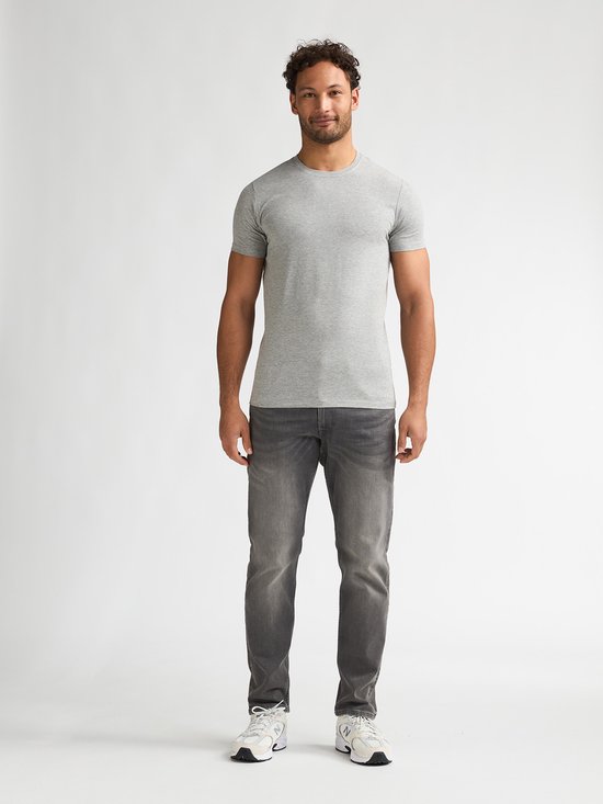 Man met krullend haar draagt een effen lichtgrijs T-shirt met ronde hals en grijze jeans.