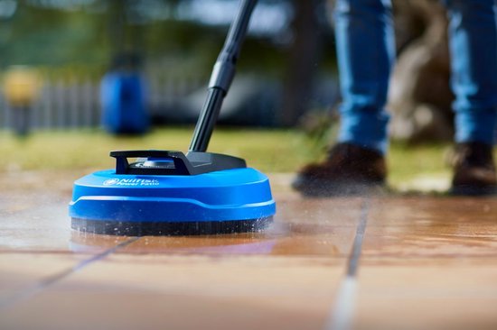 Blauwe Nilfisk terrasreiniger die water sproeit op een betegeld terras, met benen van een persoon op de achtergrond.