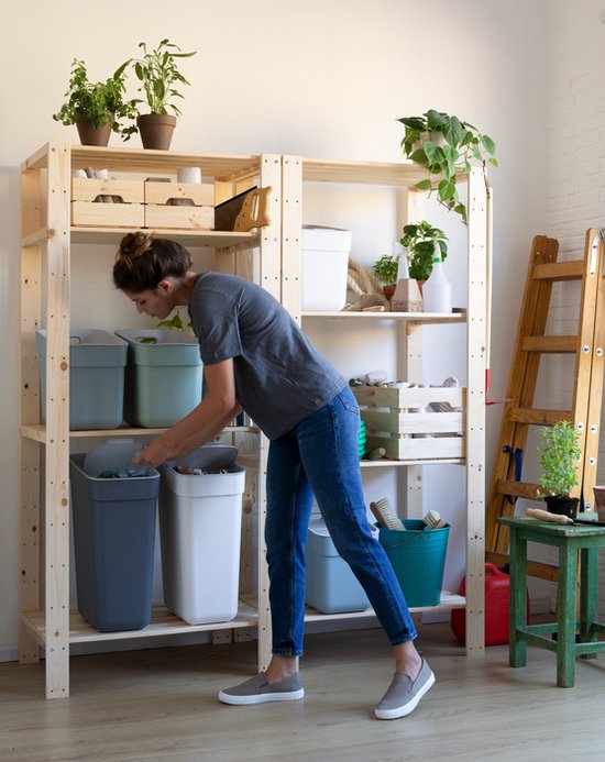 Une femme place une poubelle grise avec couvercle dans une étagère en bois. À côté d'elle se trouvent une poubelle blanche et une poubelle bleue.