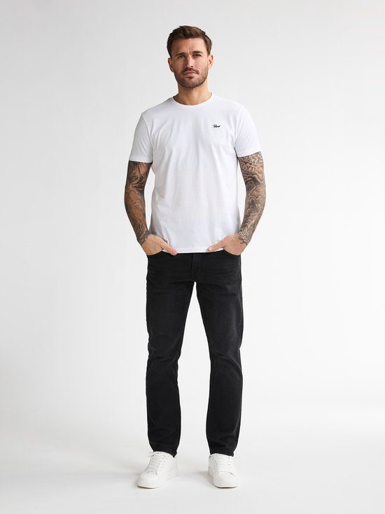 Man met tattoos op de armen in een wit T-shirt en zwarte jeans.