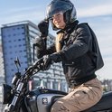Vrouw in beige motorbroek en zwarte jas op motorfiets, met helm in hand.