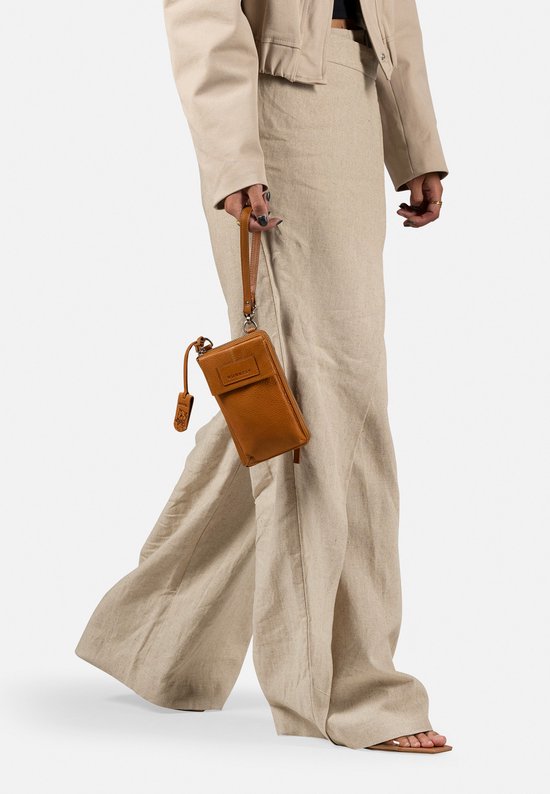 Model met cognac gekleurde telefoonportemonnee met polsbandje, gedragen bij een beige outfit.