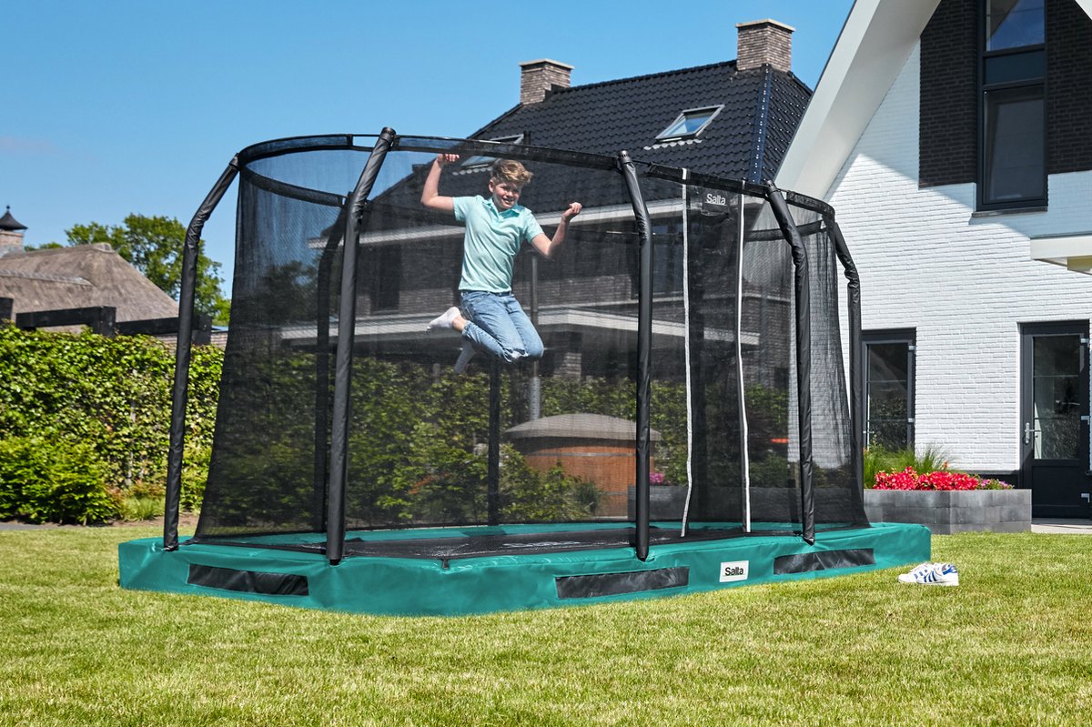 Jongen springt op een rechthoekige inground trampoline met veiligheidsnet in een gazon naast het huis