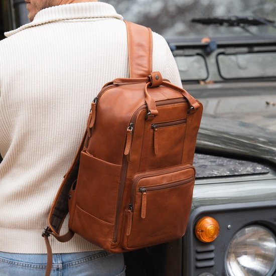 Man draagt een cognac leren rugzak, staand naast de voorkant van een donkere terreinwagen.
