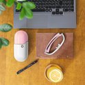 Een witte en roze capsulevormige AYO lichttherapiebril, een zwarte pen, een glas met citroenwater en een laptop op een houten bureau.