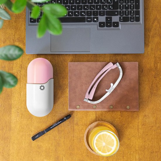 Een witte en roze capsulevormige AYO lichttherapiebril, een zwarte pen, een glas met citroenwater en een laptop op een houten bureau.