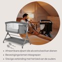 Grijze co-sleeper met afneembare zijkant, bevestigingsriemen en een moeder op de achtergrond.